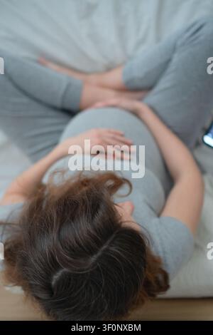 Pregnant woman sitting on white couch with hand on belly smiling happily. Expecting mother cradling stomach in warm joyful moment at home. Mom-to-be radiating love and happiness in bright living room Stock Photo