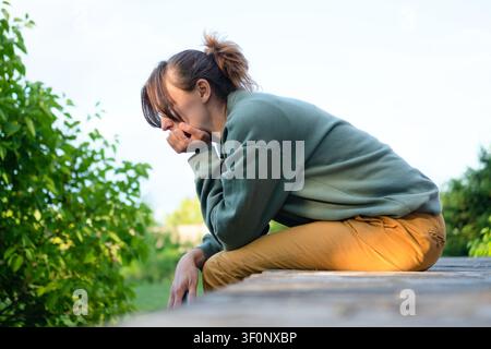 A young woman sits on a bench with her head in her hands in a green garden. The woman hiding her face presents a lonely, despair moment of emotional distress and thoughtful reflection outdoors. Stock Photo