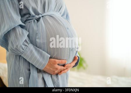 Pregnant woman back view with hands on belly in blue dress on white background. Expecting mother in relaxed confident pose at studio. Mom-to-be showing happy motherhood in stylish maternity wear Stock Photo
