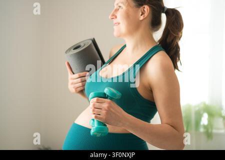 Pregnant woman in comfortable activewear side view for prenatal workout. Expecting mother wears modern maternity fitness outfit with dumbbell. Mom-to-be stays confident and stylish during exercise Stock Photo