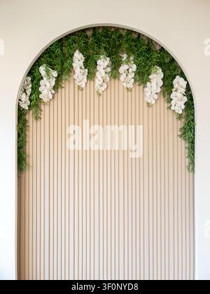 Elegant floral arch featuring cascading white orchids and lush greenery on a neutral wooden slat backdrop. Stock Photo