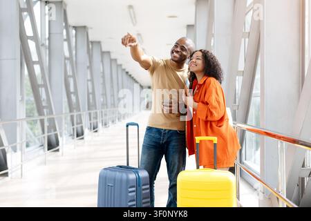Smiling young Black couple standing with suitcases in airport, pointing toward gate Stock Photo