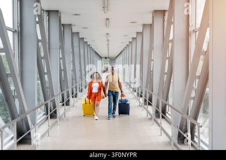 Smiling Black couple walks hand in hand through airport with colorful luggage Stock Photo
