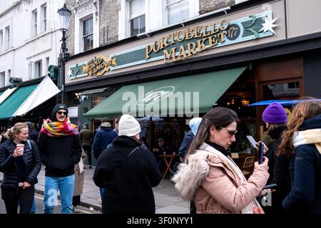 People walk and gather outside a storefront with a large sign reading The World Famous Portobello Market 177 and a green awning at Portobello Road Mar Stock Photo