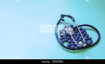Fresh organic blueberries arranged in a heart shape with a black medical stethoscope on a turquoise background. Concept of healthy eating and cardiac Stock Photo