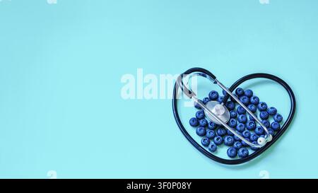 Fresh organic blueberries arranged in a heart shape with a black medical stethoscope on a turquoise background. Concept of healthy eating and cardiac Stock Photo