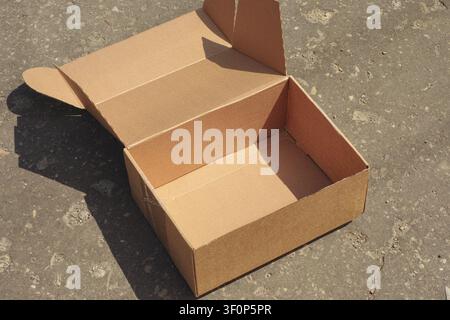 An empty open brown cardboard box sitting on a textured asphalt surface outdoors under bright sunlight. Stock Photo