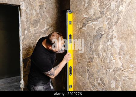 Builder checking the evenness of marble tiles with a level tool, ensuring precision and accuracy during renovation work on a wall Stock Photo