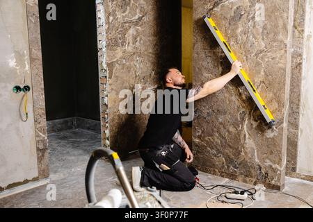 Builder checking the evenness of marble tiles with a level tool, ensuring precision and accuracy during renovation work on a wall Stock Photo