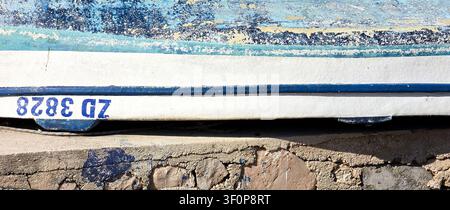 A detailed view of a distressed blue and white boat hull showing peeling paint and a visible registration number on a concrete dock surface beneath. Stock Photo