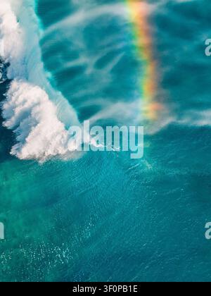 Aerial view of a powerful ocean wave breaking in turquoise water with a vibrant rainbow forming in the sea spray in Mauritius. Stock Photo