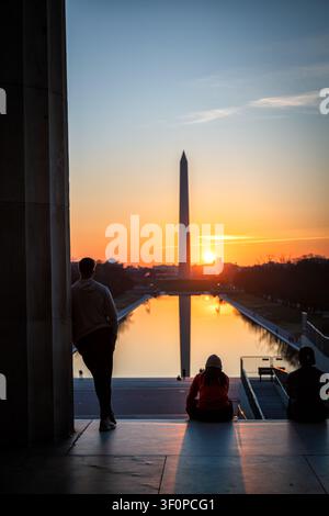 Sunrise over Washington Monument from Lincoln Memorial | WASHINGTON D.C., United States — Silhouetted visitors watch sunrise align with the Washington Monument from the Lincoln Memorial steps, its obelisk mirrored in the Reflecting Pool below. Henry Bacon designed the pool as a passive mirror extending the memorial's axis toward the Capitol, part of the 1901 McMillan Plan built atop reclaimed Potomac tidal flats. The basin stretches roughly 2,028 feet (618 m) toward the monument. Stock Photo