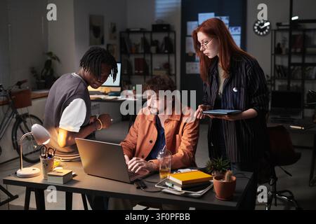 Diverse Coworkers Collaborating on Laptop in Office at Night Stock Photo