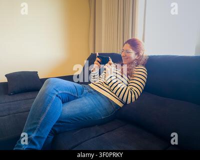 Woman smiling and using smartphone while lounging on a comfortable sofa in her living room Stock Photo