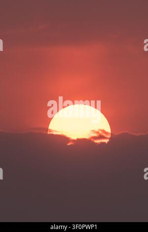 An abstract and vertical shot of a vibrant sunset with the large, golden sun partially hidden behind a horizontal band of dark clouds, with a beautifu Stock Photo