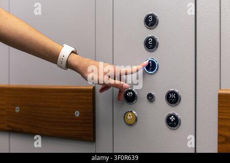 Hand pressing illuminated elevator button. A hand wearing a white smartwatch presses the illuminated number one button on a gray elevator control Stock Photo