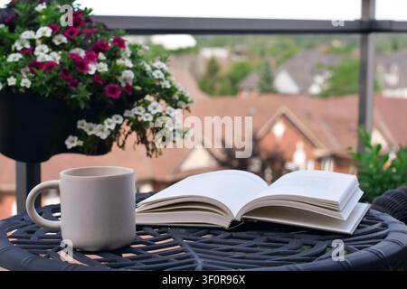 Eye-level close-up showing open book and white mug on black metal table with hanging flower basket and blurred rooftop background Stock Photo