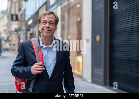 Smiling 55 year old man in a business suit walks along a modern city street with a red backpack Stock Photo