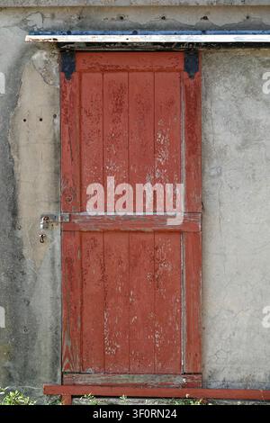 Red wooden door with vertical planks, slightly weathered, showing peeling paint. It features metal hinges and is set against a rough concrete wall. Stock Photo