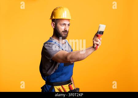 Bearded painter in safety helmet extends paintbrush forward as if preparing to paint surface. Confident male contractor poses in studio, dressed in construction overalls against yellow background. Stock Photo