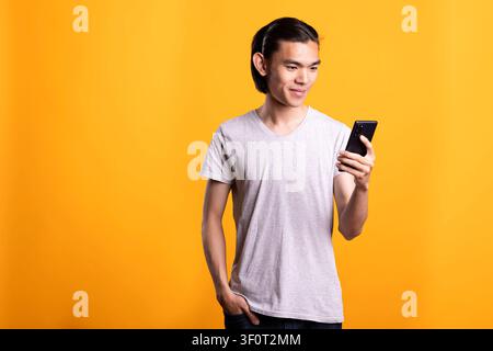 Handsome asian man in casual outfit standing with hand in pocket, using smartphone to chat with friends. Modern portrait of young male adult communicating online through mobile device and social media Stock Photo