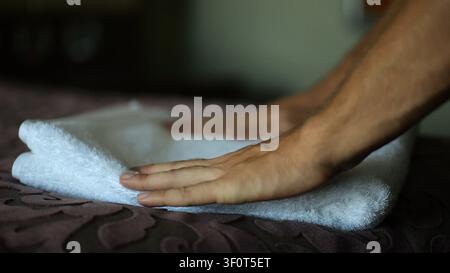 Male hands of housekeeping adjusting soft white terry towel on a neatly made bed in a modern hotel room. Service personnel preparing clean bedding and Stock Photo