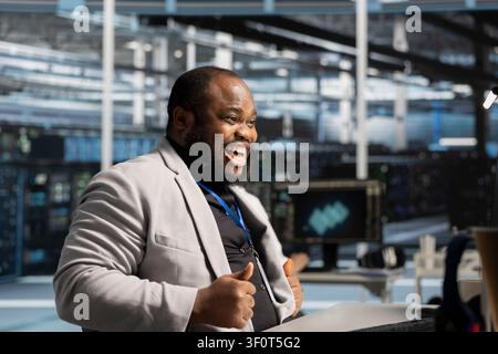 Delighted coder in server facility satisfied by improved system efficiency metrics. Ecstatic African American man working in server facility satisfied by enhanced network infrastructure performance. Stock Photo