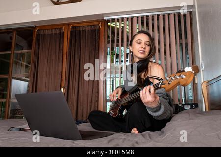 Portrait of a smiling young woman practicing electric bass guitar while wearing wireless headphones and using a laptop. Stock Photo