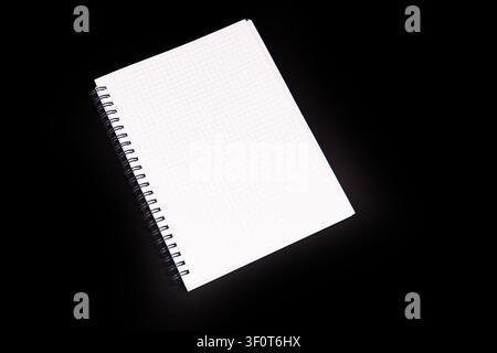 Graph paper notebook with spiral binding, positioned diagonally against black background, revealing white pages and faint grid pattern Stock Photo