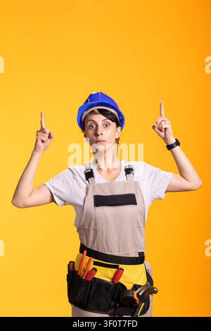Caucasian female builder gestures upward as if signaling attention to announcement or idea. Renovation worker stands against studio background portraying awareness and message delivery. Stock Photo