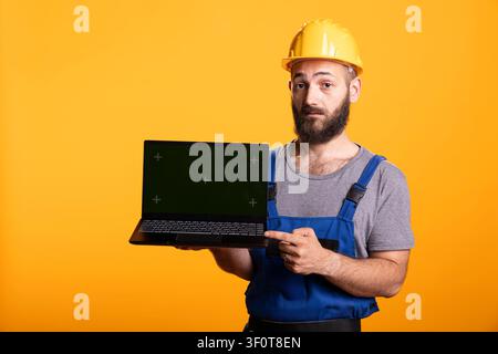 White male contractor presents open laptop displaying green screen, suitable for promotional construction content. Repairman holds device as copy space template for renovation or DIY advertisements. Stock Photo