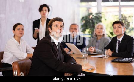Concentrated young adult old office workers sitting around the table and looking at screen while Latin female using projector remote control in office Stock Photo