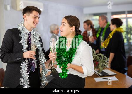 Cheerful young man and woman standing with glasses of champagne during corporate party with colleagues in meeting room Stock Photo