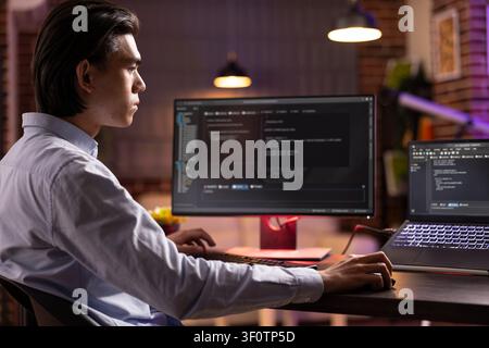 Young male freelancer sits in home office during evening hours, analyzing and debugging code on laptop and desktop PC. Asian entrepreneur developing web applications for online company. Stock Photo