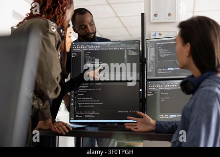 Multiethnic programmers collaborate, reviewing code on computer screens to develop functional website. African American and Asian colleagues analyze HTML scripts across multiple monitors on desk. Stock Photo