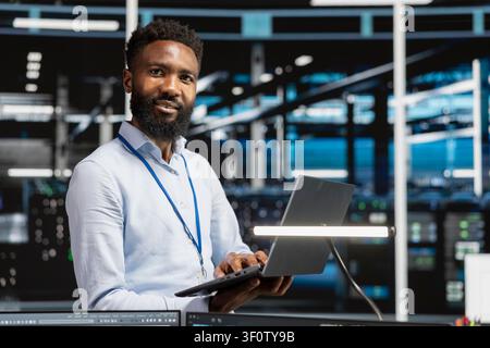 Cheerful data center engineer monitors system performance on a laptop, identifying bottlenecks and troubleshooting server hub errors. Stock Photo
