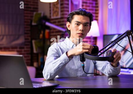 Asian man sits at desk with microphone on tripod, holding listening device and filming professional podcast episode. Male vlogger promoting electronics and sharing product reviews with online audience Stock Photo