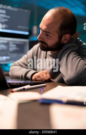 Stressed male developer leaning on hand with pensive expression, struggling with challenging programming project. Exhausted programmer thoughtfully sits at desk, looking at laptop during night shift. Stock Photo