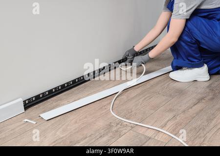 Worker installing a white plastic baseboard with a cable channel on a wooden floor, home renovation and interior design, the person wears blue overall Stock Photo