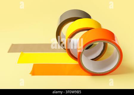 Three rolls of colorful adhesive tape in orange, yellow, and gray with strips unrolled on a light yellow background Stock Photo