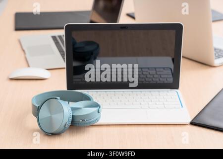 Light blue wireless headphones resting on a white laptop keyboard on a wooden desk with Other devices, Other laptops and a computer mouse are visible Stock Photo