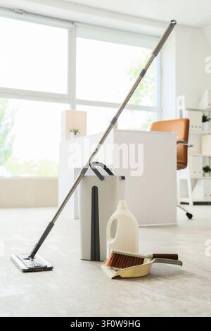 Cleaning supplies including a mop, bucket, detergent, brush, and dustpan on a light floor in an office setting Stock Photo