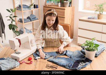 Young woman cutting denim jeans with scissors at a sewing machine on a wooden table fashion design and upcycling, this scene fashion design upcycling Stock Photo