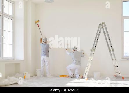 Team of professional painters in uniform painting white wall in empty room with paint rollers. Stock Photo