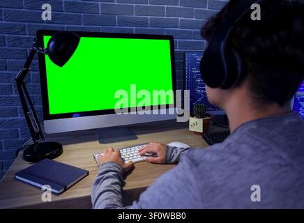 Young man wearing headphones working on computer with green screen monitor and coding on second screen at night, a second monitor shows lines of code, Stock Photo