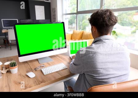 Man working on desktop computer and laptop with green screens in a modern office setting Stock Photo