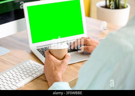 Person holding a disposable coffee cup while typing on a laptop with a green screen at a wooden desk, Modern work environment with a wooden desk, emph Stock Photo