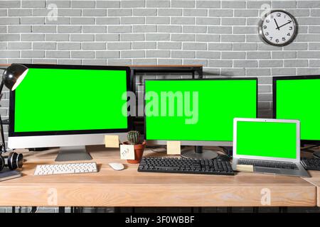 Multiple computer monitors and a laptop with green screens on a wooden desk in a modern office setting Stock Photo