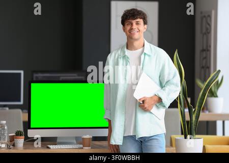 Smiling young man holding a laptop standing next to a computer with a green screen in a modern office Stock Photo