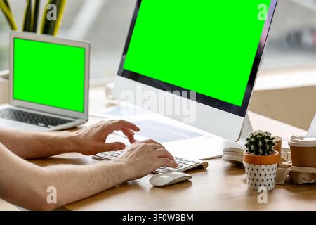 Person typing on a keyboard with a green screen desktop monitor and laptop on a wooden desk in an office setting Stock Photo